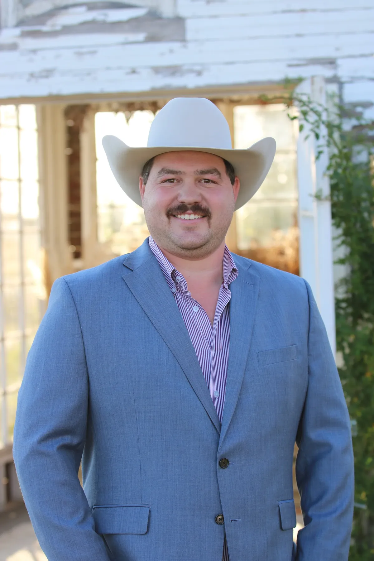 Matt Zajic in a light blue sport coat and white cowboy hat, standing in front of a weathered white farmhouse